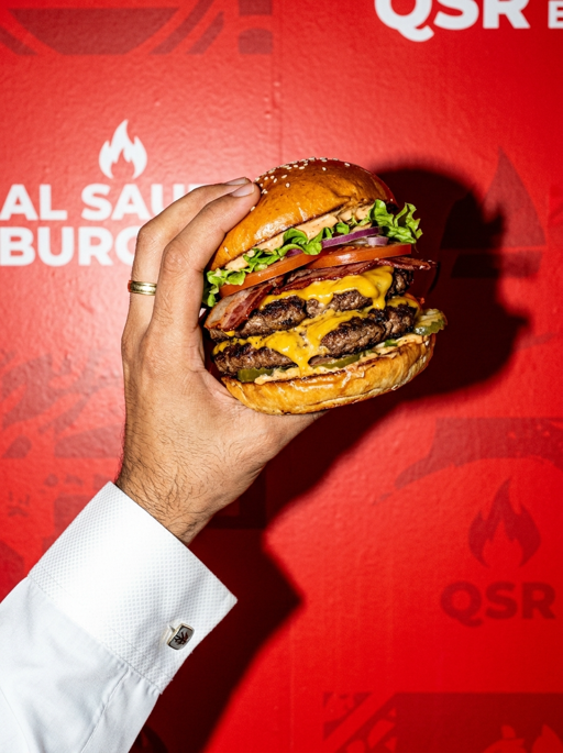 AI product photography scene — food hand holding loaded burger red wall | rawbit AI product photography scene — food hand holding loaded burger red wall | rawbit